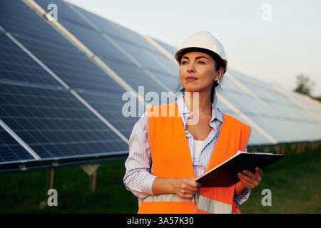 Checking the data, with notepad. Female worker engineer in uniform is near solar panels. Stock Photo