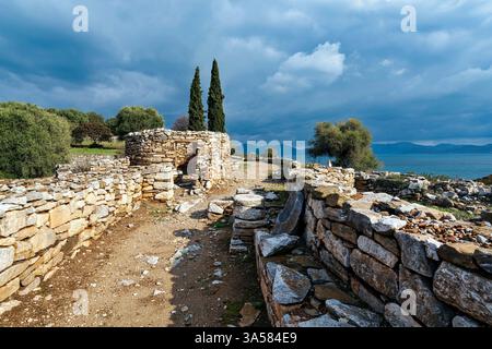 A historic stone pathway winding through the ruins of Ramnous, Greece, with ancient walls, cypress trees, and a view of the Aegean Sea. Stock Photo