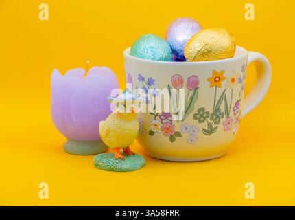 A festive Easter still life with a floral mug of chocolate eggs, a yellow chick figurine, and a purple candle against a vibrant yellow background Stock Photo