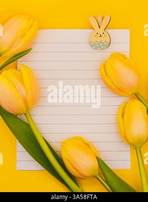 A blank notebook sheet clipped with a bunny clip, surrounded by soft beige-yellow tulips on a vibrant yellow background. Perfect for Easter cards Stock Photo
