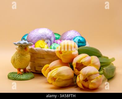 Easter still life with a decorative chick, a basket of colourful chocolate eggs, and fresh yellow tulips on a soft beige background. Stock Photo