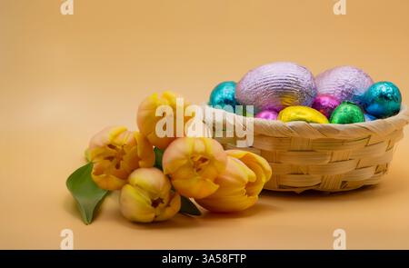 Easter still life with a basket of colourful chocolate eggs and fresh yellow tulips on a soft beige background, perfect for festive designs. Stock Photo