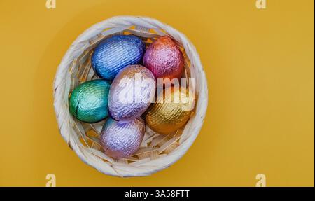 Easter still life with a wicker basket filled with colourful eggs on a vibrant yellow background, perfect for festive designs and decorations. Stock Photo