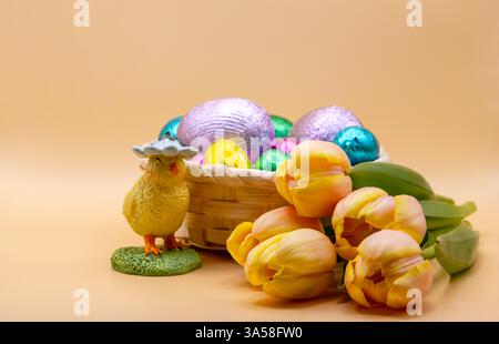 Easter still life with a decorative chick, a basket of colourful chocolate eggs, and fresh yellow tulips on a soft beige background. Stock Photo