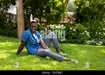 Smiling man wearing bunny ears sitting on grass in sunny garden Stock Photo