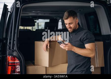 Using the smartphone. Delivery man or loader is near the car with a box. Stock Photo