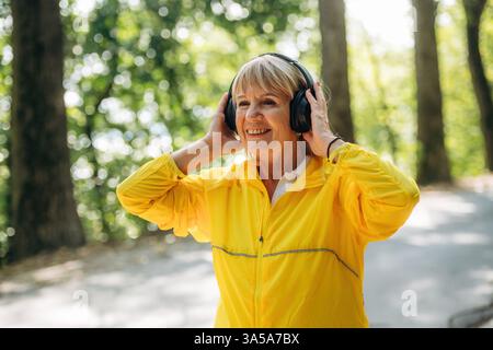 Senior woman in yellow sport coat is outdoors with headphones. Stock Photo