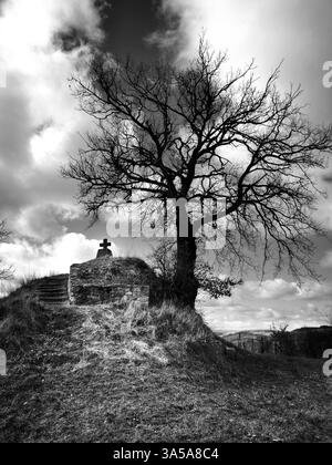 A solitary tree stands on a hill, framed by dark clouds. A cross can be seen nearby, hinting at a place of reflection. Stock Photo