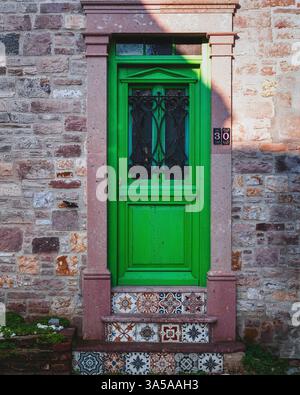 Bright green wooden door with black iron bars and colorful tiled steps set in a stone wall in a historic Mediterranean village. Stock Photo