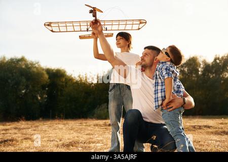 Sitting, side view. Family of father, son and daughter are playing with wooden toy plane on the field. Stock Photo