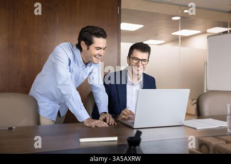 Two men colleagues laughing, look at laptop screen, accomplishing task Stock Photo