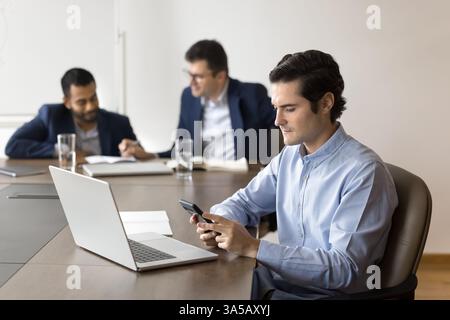 Young focused businessman using smartphone sit at desk with laptop Stock Photo