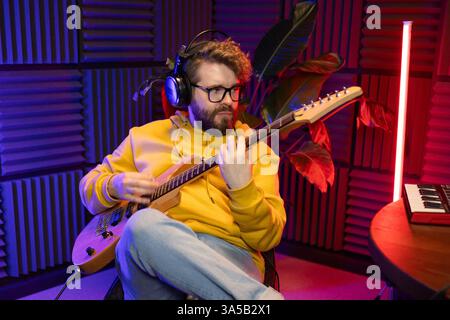 A focused male musician fine-tunes a guitar against the backdrop of a digital sound editing setup in a contemporary recording studio environment. Stock Photo