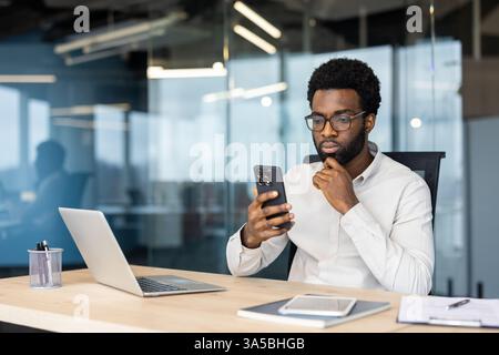 A serious, young professional man in glasses checks his smartphone while sitting at his desk in a modern office. Stock Photo