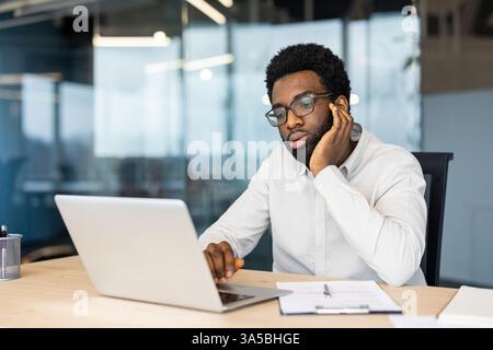 A young professional contemplates at his desk in a modern office, with a laptop and documents beside him. Stock Photo