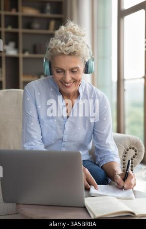 Older female wearing headphones communicating by videocall via notebook handwriting Stock Photo