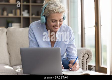 Elderly lady in headset watching webinar on notebook taking notes Stock Photo