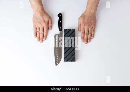 on white background on the table there lies a large knife for sharpening and a black grooved sharpening stone on the first floor, men's hands lie next Stock Photo