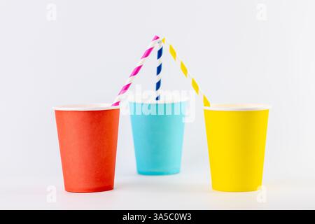 on white background there are three paper cups next to each other yellow red and blue with matching paper cocktail straws inside disposable tableware Stock Photo