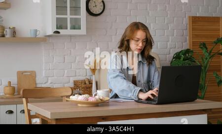 Engrossed in her work, a woman writes in a notebook while using a laptop in a bright kitchen. This image reflects the flexibility and comfort of Stock Photo