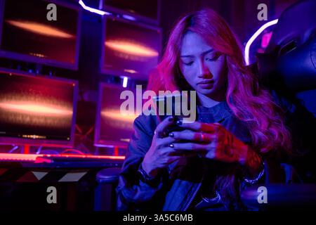 Asian woman in front of a computer monitor display using mobile phone, neon light, Hacker wear black Stock Photo