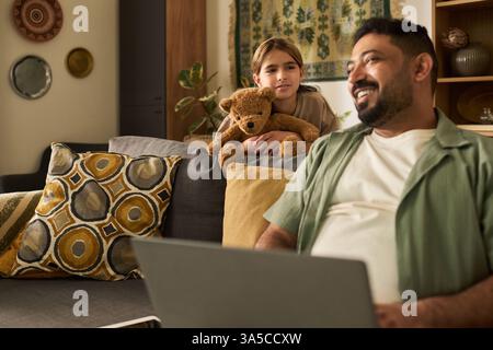 Father smiling while using laptop, daughter holding teddy bear sitting beside him on couch in cozy living room with decorative pillows and art pieces hanging on wall Stock Photo