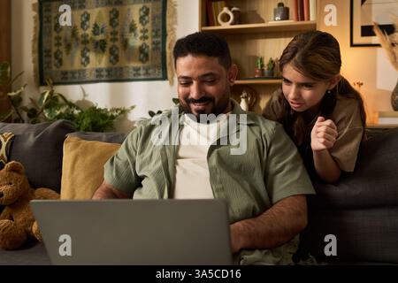 Man smiling while using laptop can be seen with woman leaning closer in cozy room with shelves and plant adds a warm touch. Wall decor and cushion complete the setting Stock Photo