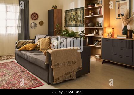 Living room interior showcasing eclectic mix of bohemian decor with cozy couch, patterned rug, and shelf of books. Soft lighting from lamp creating warm and inviting atmosphere Stock Photo