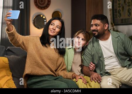 Woman, man, and girl sitting on sofa, taking selfie with bright smiles on their faces, showing warmth and closeness in indoor setting Stock Photo