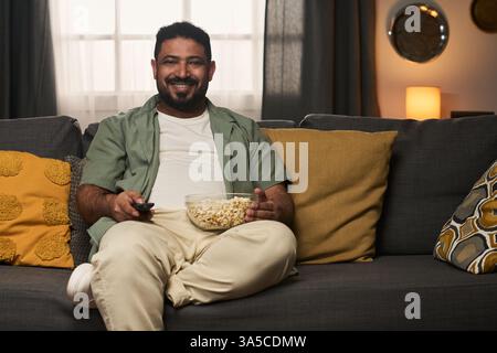 Man relaxing on couch holding bowl of popcorn with relaxed expression, smiling warmly with cozy living room setup around. Background includes soft lighting and decorative pillows Stock Photo