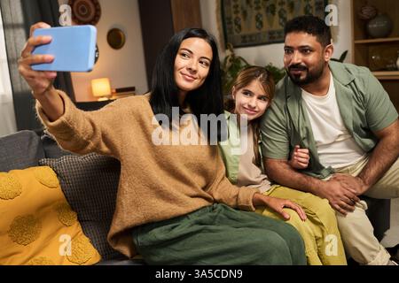 Family enjoying time together sitting on comfortable couch. Smiling while taking selfie with smartphone in cozy living room Stock Photo