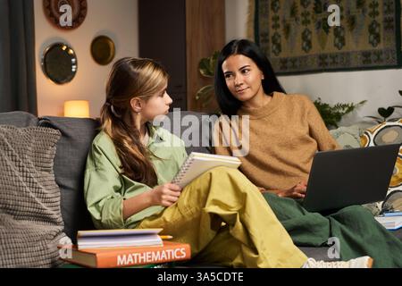 Two women engaging in study session on couch, one holding notebook and writing while other looks on supportively Stock Photo