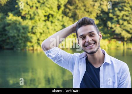 Portrait of contemplative light brown haired young man wearing white shirt beside picturesque river or lake, smiling to camera Stock Photo