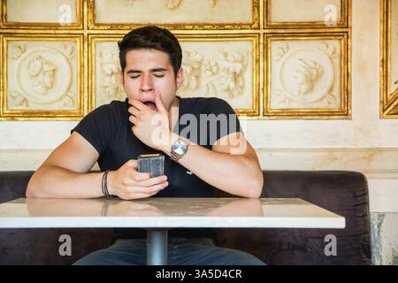 Young handsome man bored, yawning while sitting in elegant cafeteria or restaurant using cell phone to send text message Stock Photo
