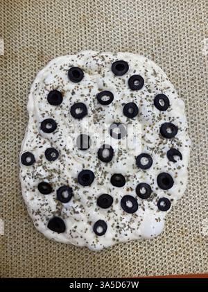 A raw pizza dough topped with sliced black olives and sprinkled with herbs, placed on a baking sheet. Stock Photo