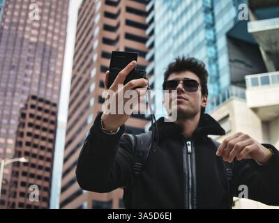 Young dark haired man standing in modern city setting, using smartphone to do videochat, or send message Stock Photo