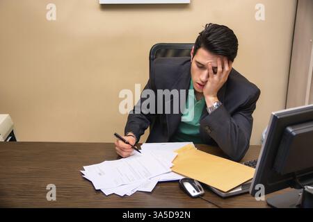 Tired bored young businessman sitting at his desk in front of his computer yawning, with his chin resting on his hand and eyes closed, in his office Stock Photo