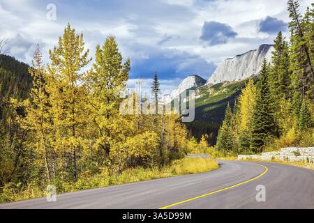 The road is among autumn dense evergreen and deciduous forests. The dramatic storm clouds over the Rocky Mountains Stock Photo