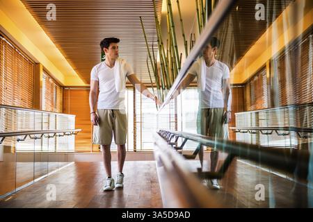 Low Angle View Full Length of Young Man with Towel Over Shoulder Standing in Bright Modern Hallway Holding on to Hand Rail Stock Photo