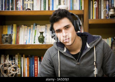 Attractive young man sitting listening to music on a set of stereo headphones in his study at home in front of a bookcase filled with books Stock Photo