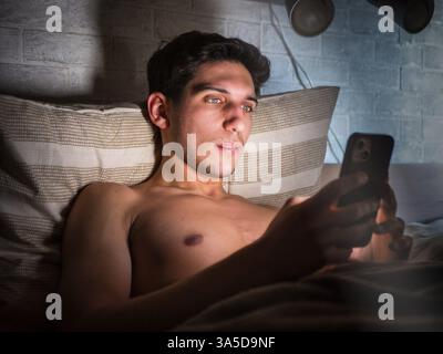 Handsome young man in bed typing on cell phone, sending text message or dialing number Stock Photo