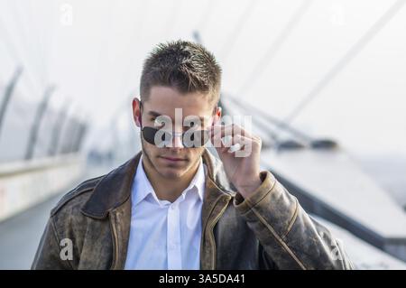 Handsome trendy young man standing on a sidewalk, wearing sunglasses and a fashionable leather jacket, in a relaxed confident pose looking at camera o Stock Photo