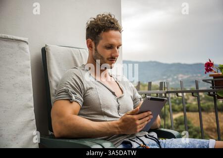 Handsome trendy man wearing t-shirt sitting and working, looking down at a tablet computer that he is holding, outdoor on house terrace Stock Photo
