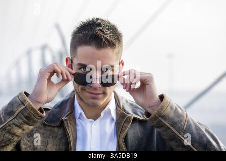 Handsome trendy young man standing on a sidewalk wearing sunglasses and a fashionable leather jacket in a relaxed confident pose looking at camera ove Stock Photo