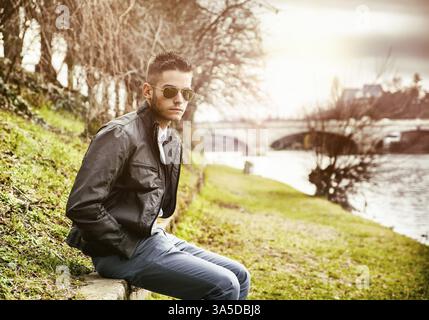 Three-quarter length of contemplative light brown haired young man wearing grey jacket and denim jeans sitting on wall beside picturesque river in Tur Stock Photo