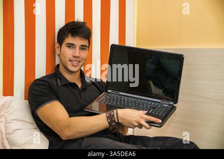 Attractive Young Man with Serious Expression Reclining Comfortably, Showing Laptop on Bed Working on his Start-up Business - Young Male College or Uni Stock Photo