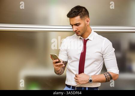 Handsome young businessman in his office typing and using cell phone, looking down, serious Stock Photo