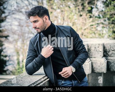 One handsome young man in urban setting in modern city, sitting, wearing black leather jacket and jeans Stock Photo