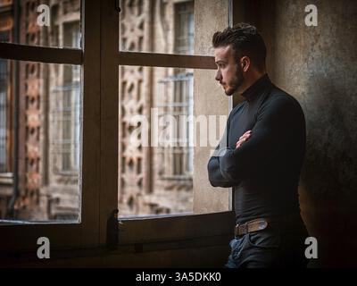 A man standing in front of a window with his arms crossed arms. Photo of a confident man with crossed arms standing in front of a window Stock Photo