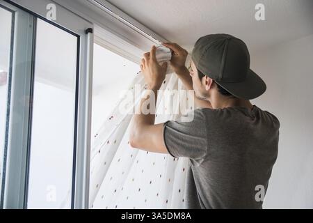 Young handsome man or handyman installing curtains over window at home, doing renovation work Stock Photo